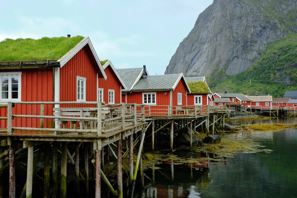 Row of red wooden houses on stilts along the shoreline with a mountain in the background.