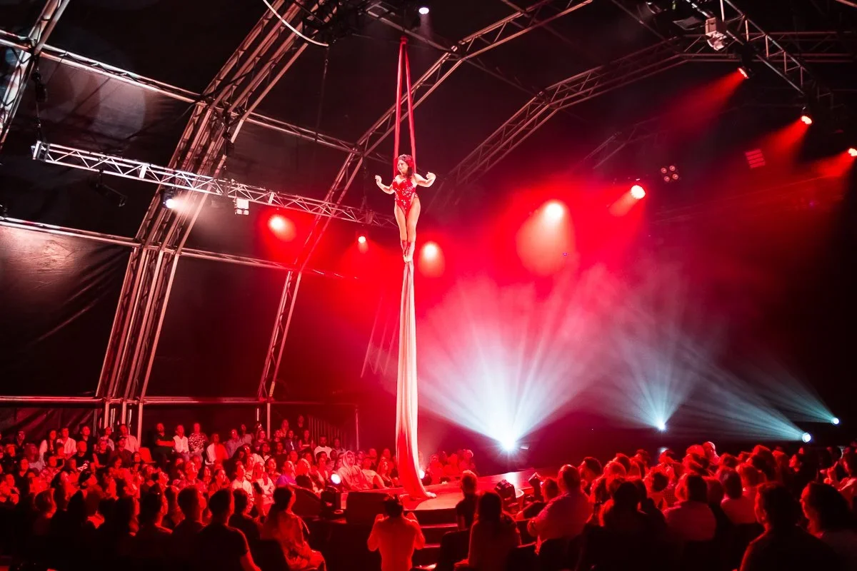 An aerial silk performer in a red costume performing in a circus tent with an audience watching, illuminated by red stage lighting.