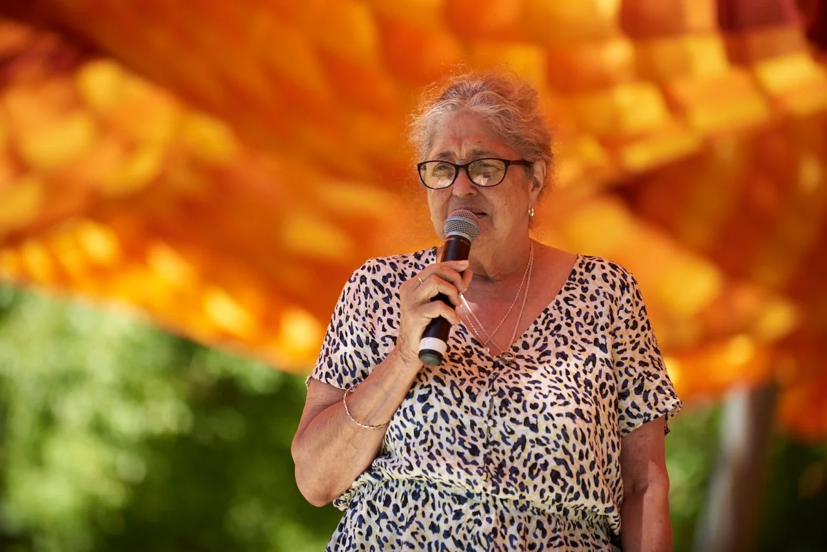 An elderly woman wearing glasses and a leopard print dress speaking into a microphone outdoors, with a blurred orange and yellow background.