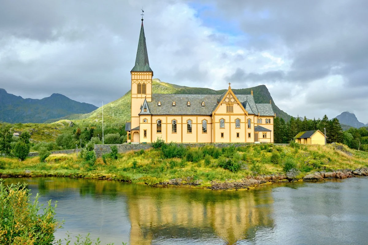 A yellow church with a tall steeple situated on a grassy hill near a body of water, with mountains and cloudy sky in the background.