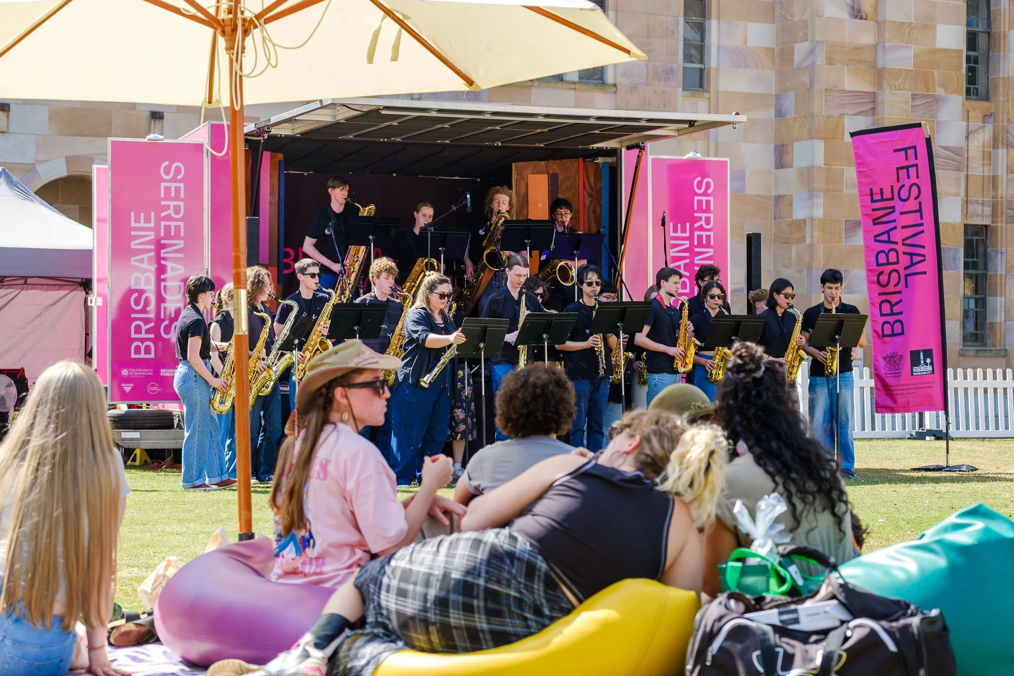 A live outdoor jazz band performs on a stage at the Brisbane Festival during daytime, with audience members sitting on the grass and some in kayaks. The stage has pink banners with event branding, and the backdrop displays a large beige brick buildin