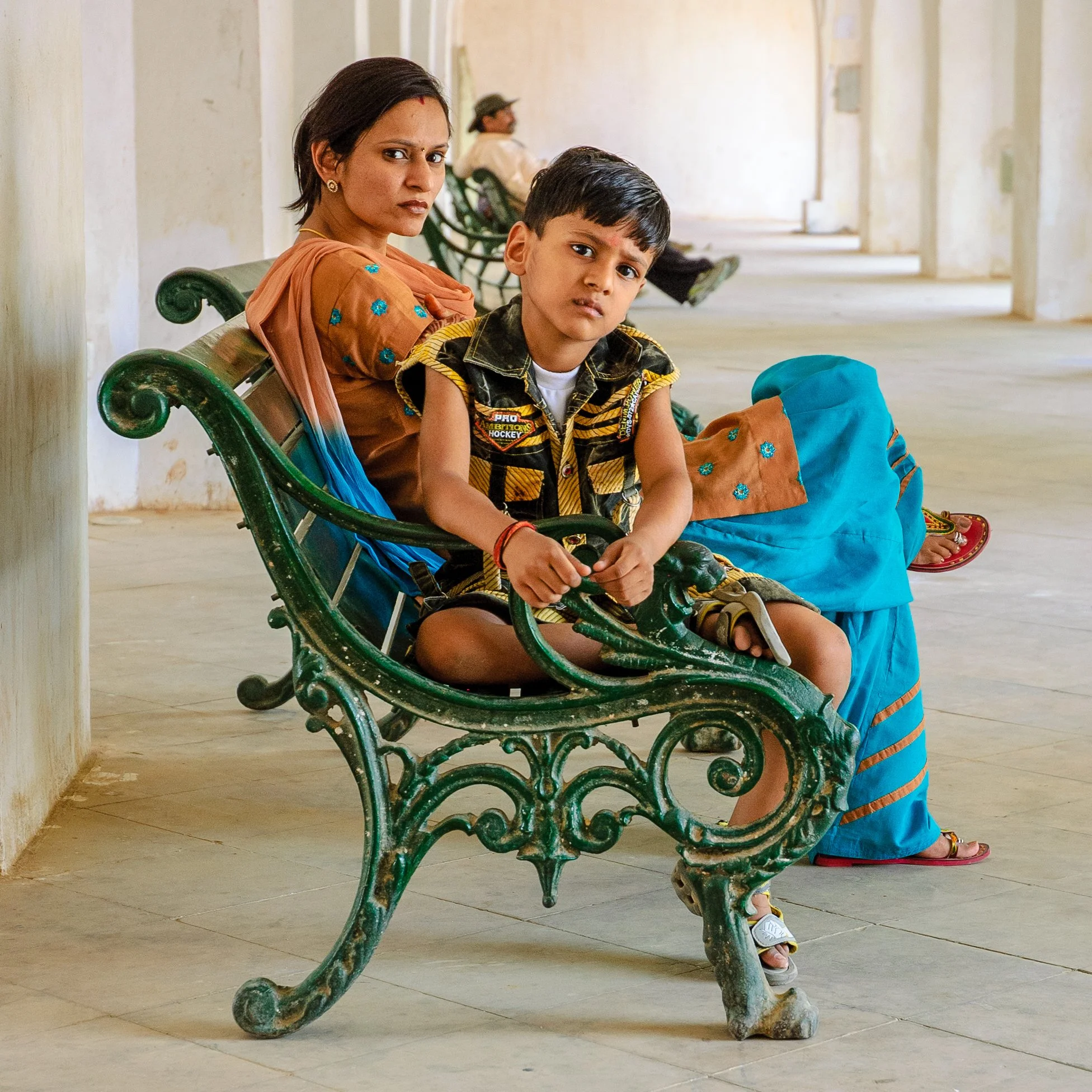 A woman and a young boy sitting on a green ornate bench in a spacious, minimally decorated corridor. The woman looks towards the camera with a serious expression, while the boy, holding the bench's armrest, looks slightly puzzled. The woman wears tra