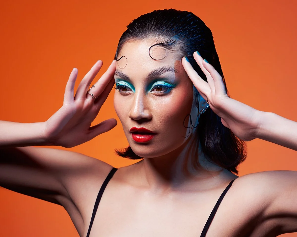 A woman with bold makeup, including blue eyeshadow and red lipstick, posing against an orange background.