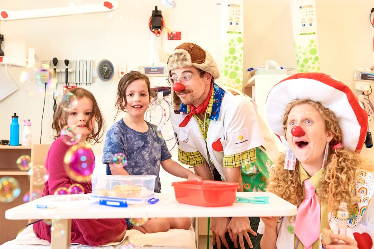 Two young girls and two clowns in a hospital room, with the girls smiling and bubbles floating around.