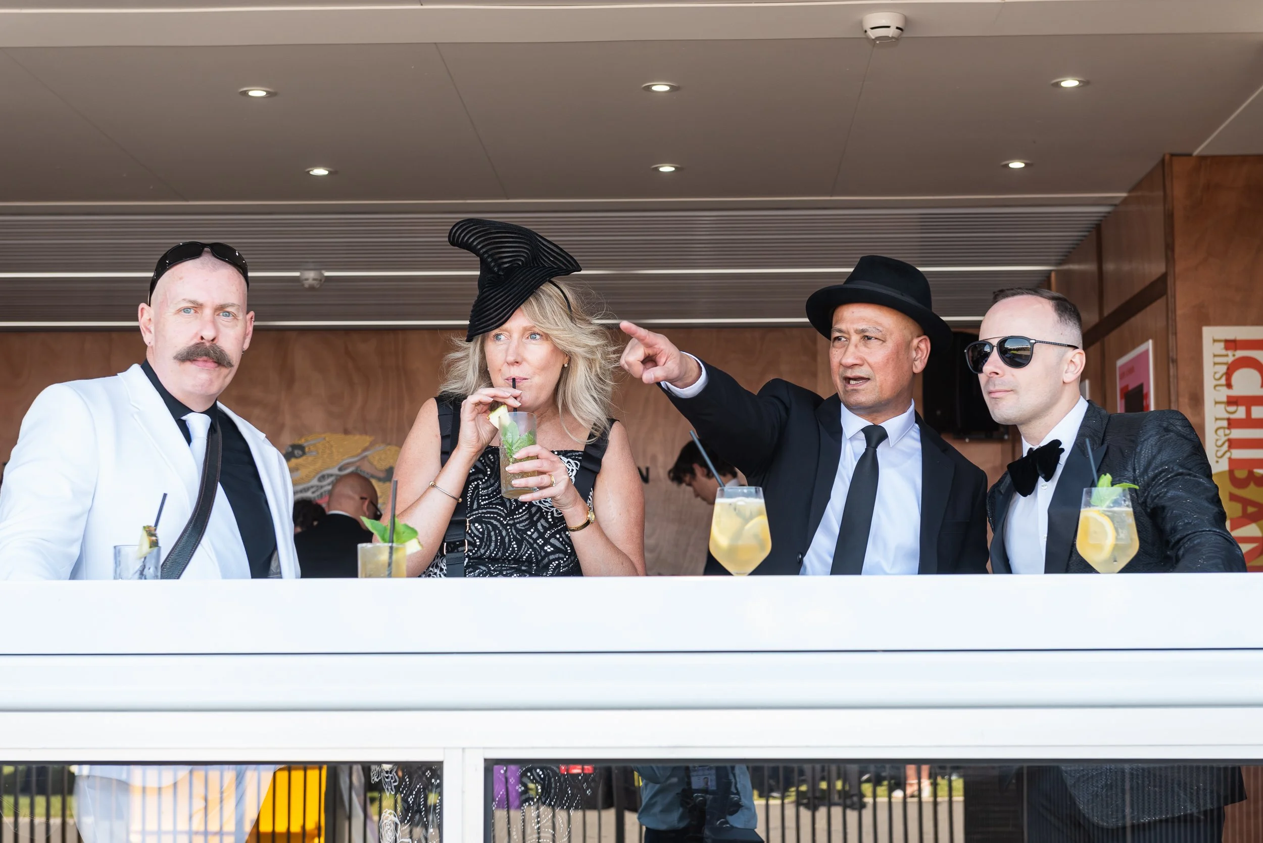 Four well-dressed people standing together at a social event, three men and one woman, with drinks. The woman is wearing a black hat with a large brim, and the man next to her is wearing a black fedora hat and pointing. The other two men are wearing 
