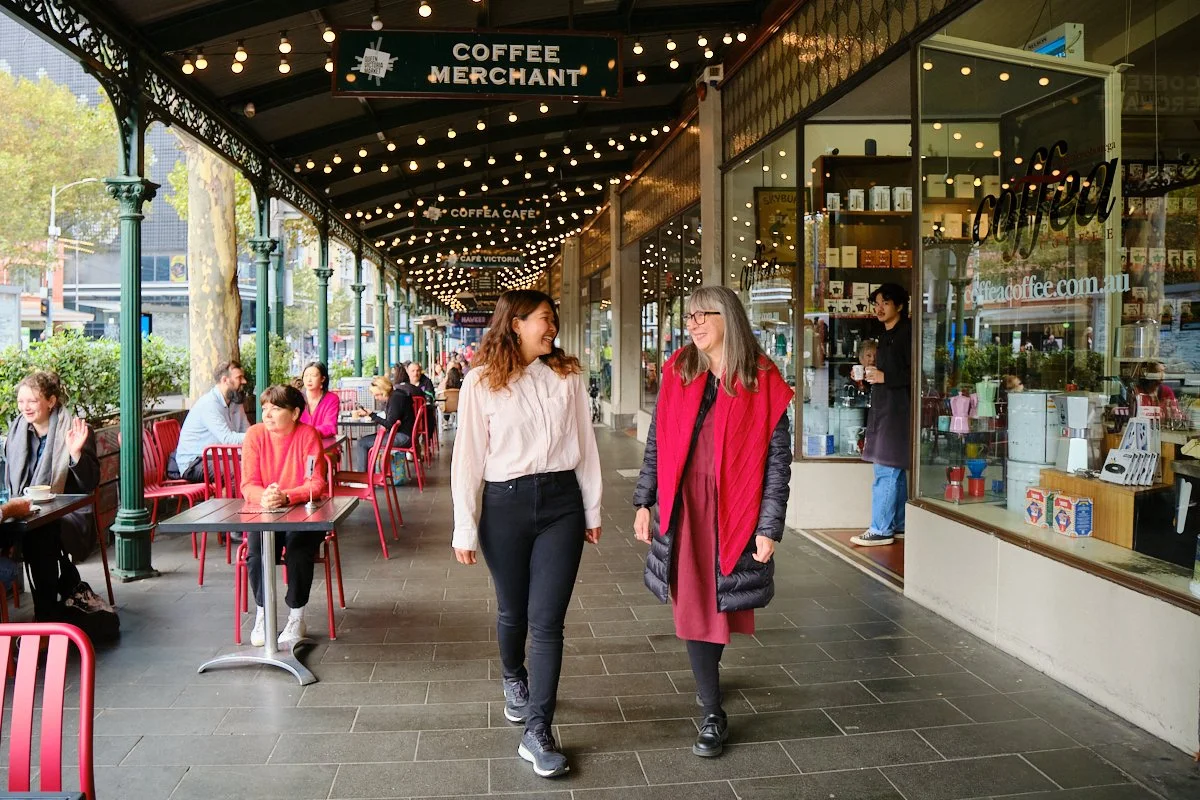 Two women are walking and talking outside a coffee shop named 'Coffee Merchant'. The shop features a glass display window with coffee products and a sign overhead. The patio has red chairs and tables, with other patrons seated, and string lights hang
