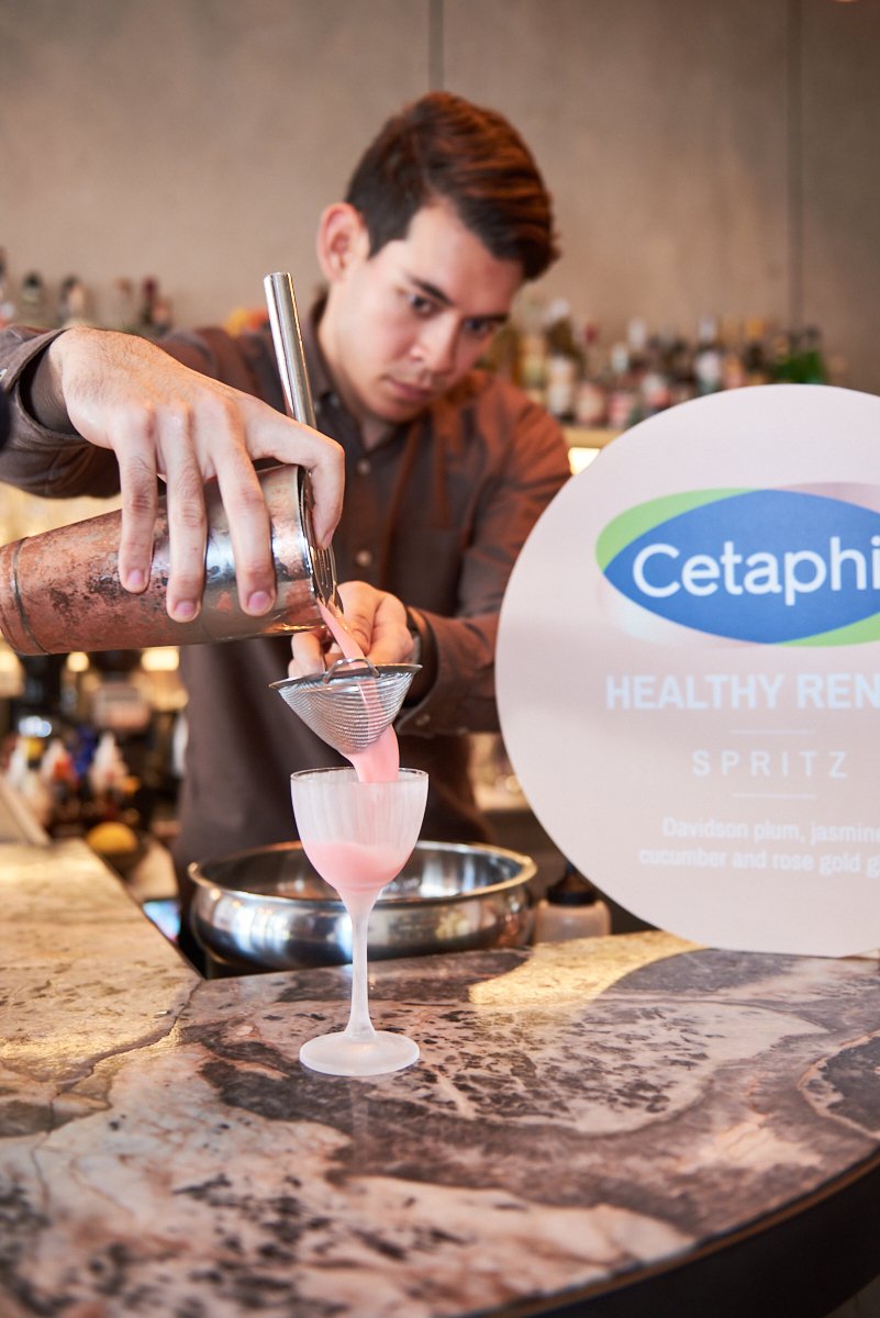 A bartender pouring a pink cocktail through a strainer into a frosted glass at a bar, with a sign in the background advertising Cetaphil healthy renewal spritz with ingredients listed.
