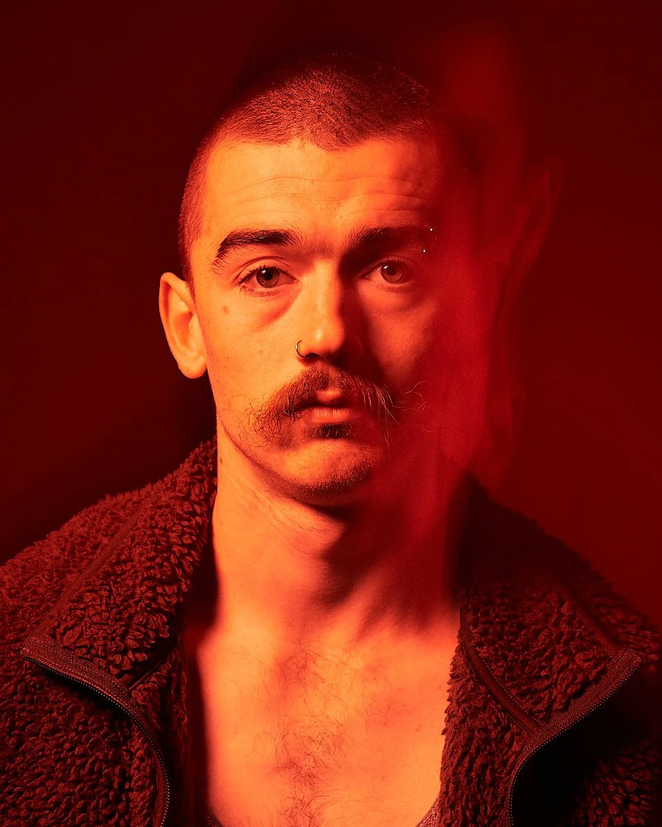 A man with short hair, mustache, and septum piercing, wearing a textured jacket, under red lighting.