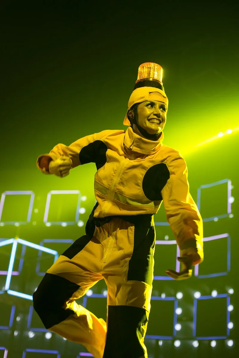 Performer in yellow and black costume with a yellow hat and a yellow hat with a light on top, smiling and looking off to the side, on a brightly lit stage with digital screens in the background.