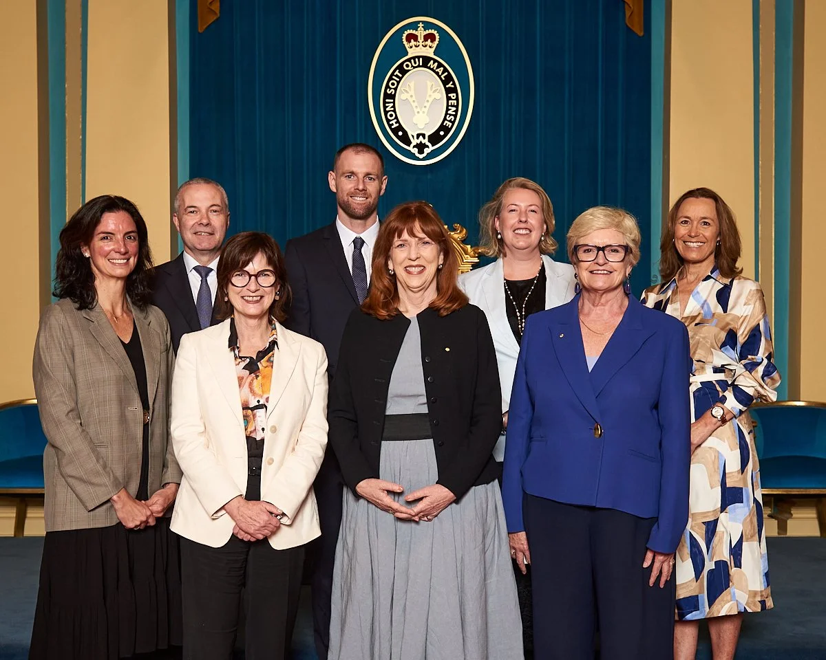 Group of eight professionally dressed women and men standing in front of a blue curtain with a crest, in a formal setting.