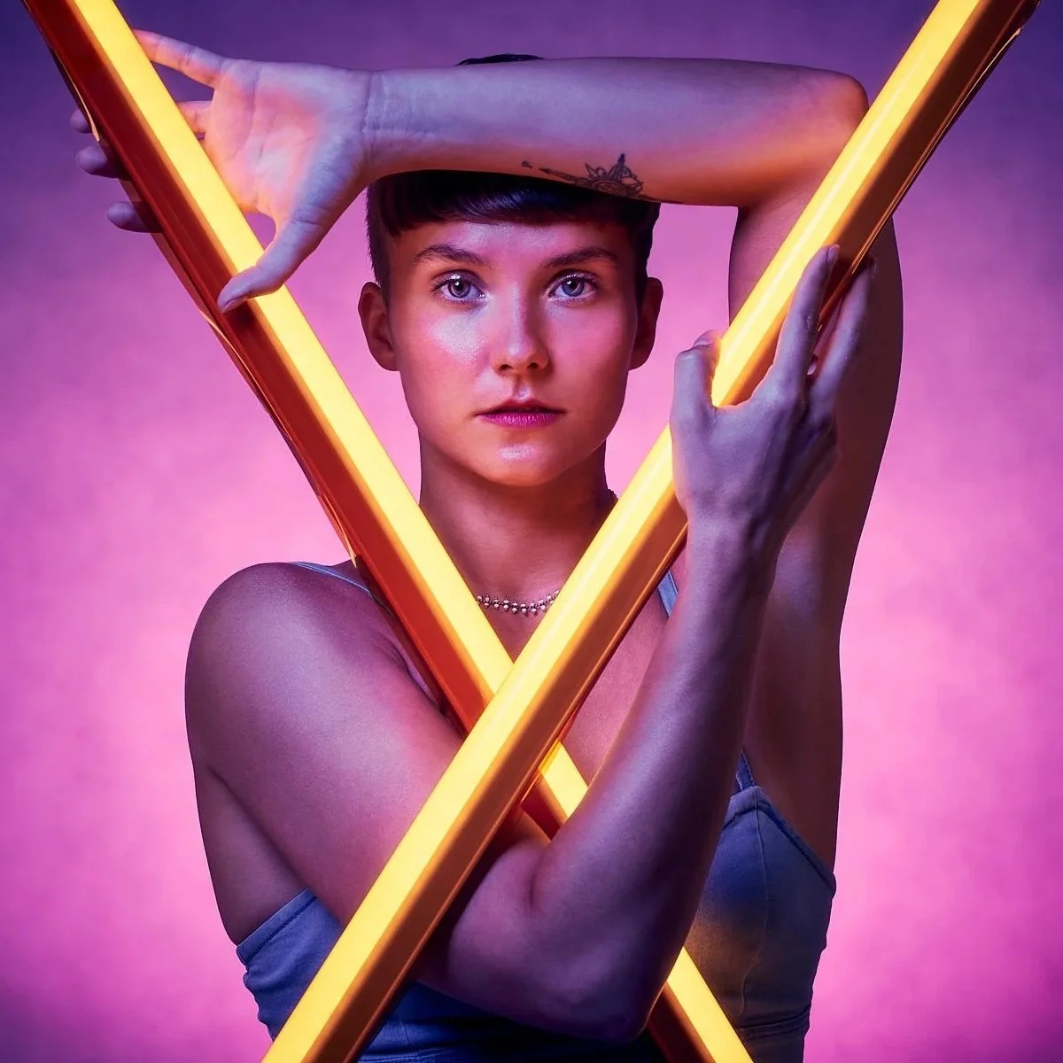 A woman with short dark hair and blue eyes holds two glowing neon yellow tubes creating a triangle shape around her face. She has a tattoo on her wrist and wears a necklace, with a pinkish-purple background.