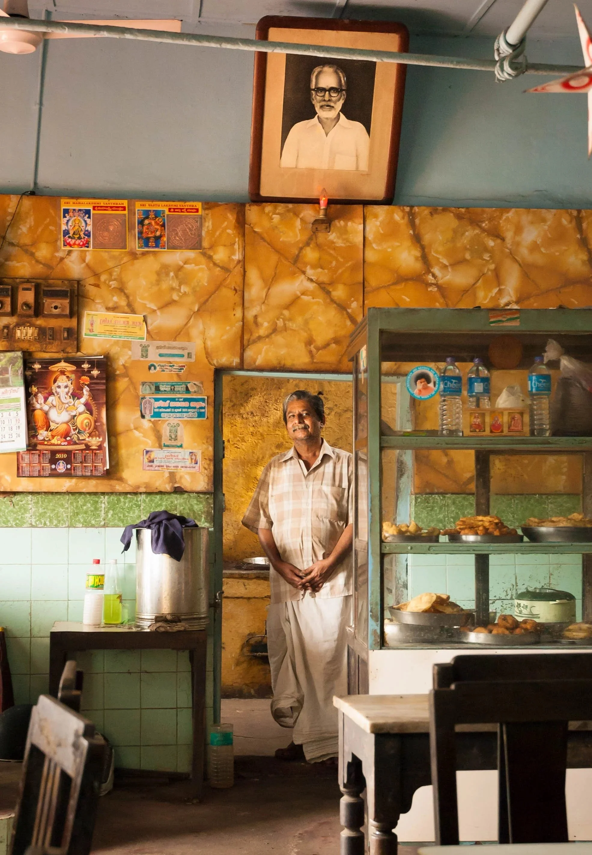 A man standing inside a small restaurant or shop, with a glass display case filled with food and water bottles, and a wall decorated with various pictures, posters, and a framed black and white portrait of a man with glasses above.