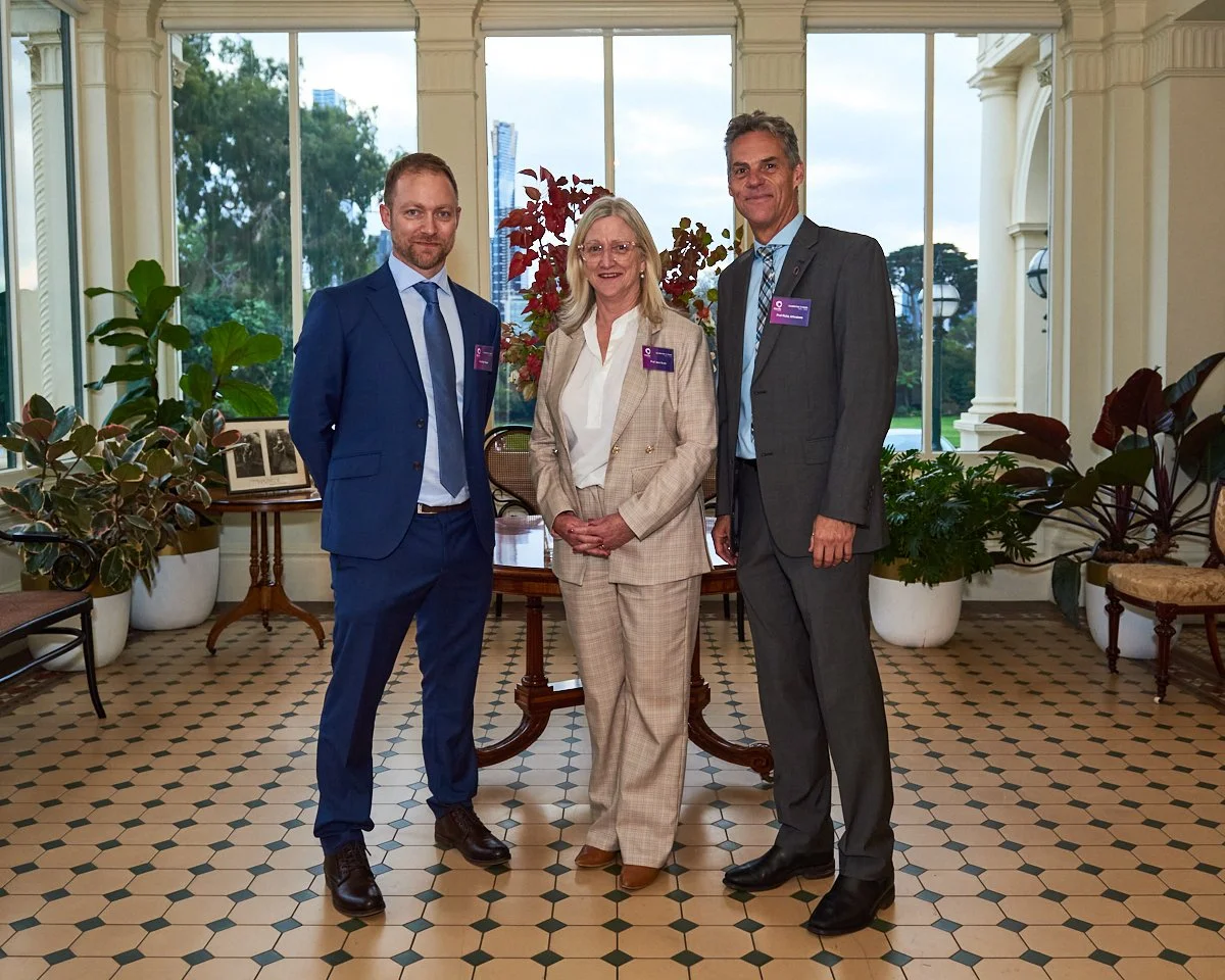 Three professionally dressed individuals, two men and one woman, standing in a formal setting with large windows and decorated with plants, smiling and facing the camera.