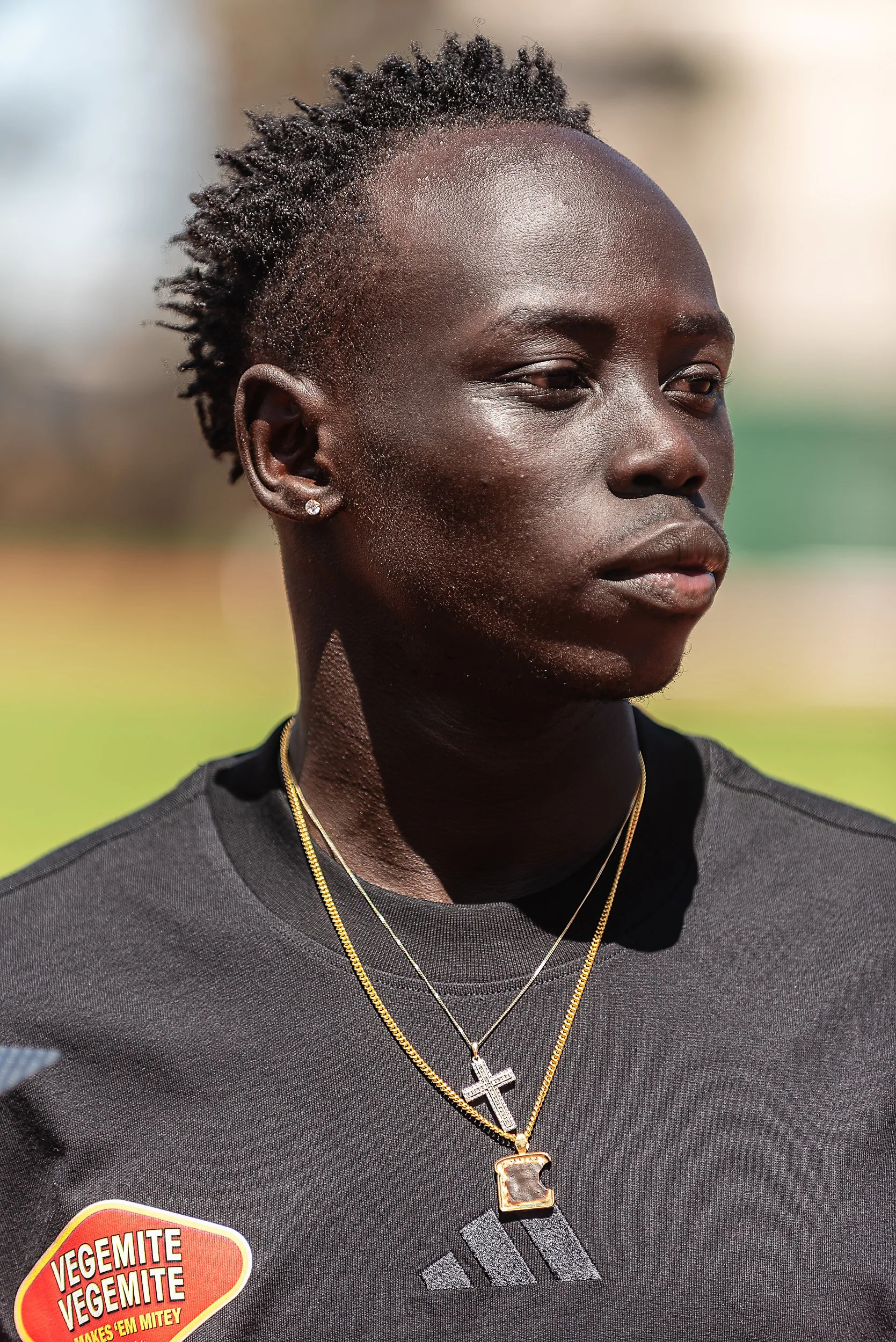 Close-up of a young person with dark skin, short curly hair, wearing earrings, and two gold necklaces with cross pendants, outdoors with a blurred background.