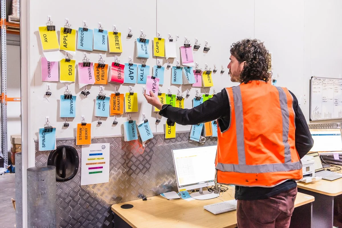 A man in an orange safety vest looking at a wall with colorful labeled cards attached by clips, possibly in a warehouse or storage facility.