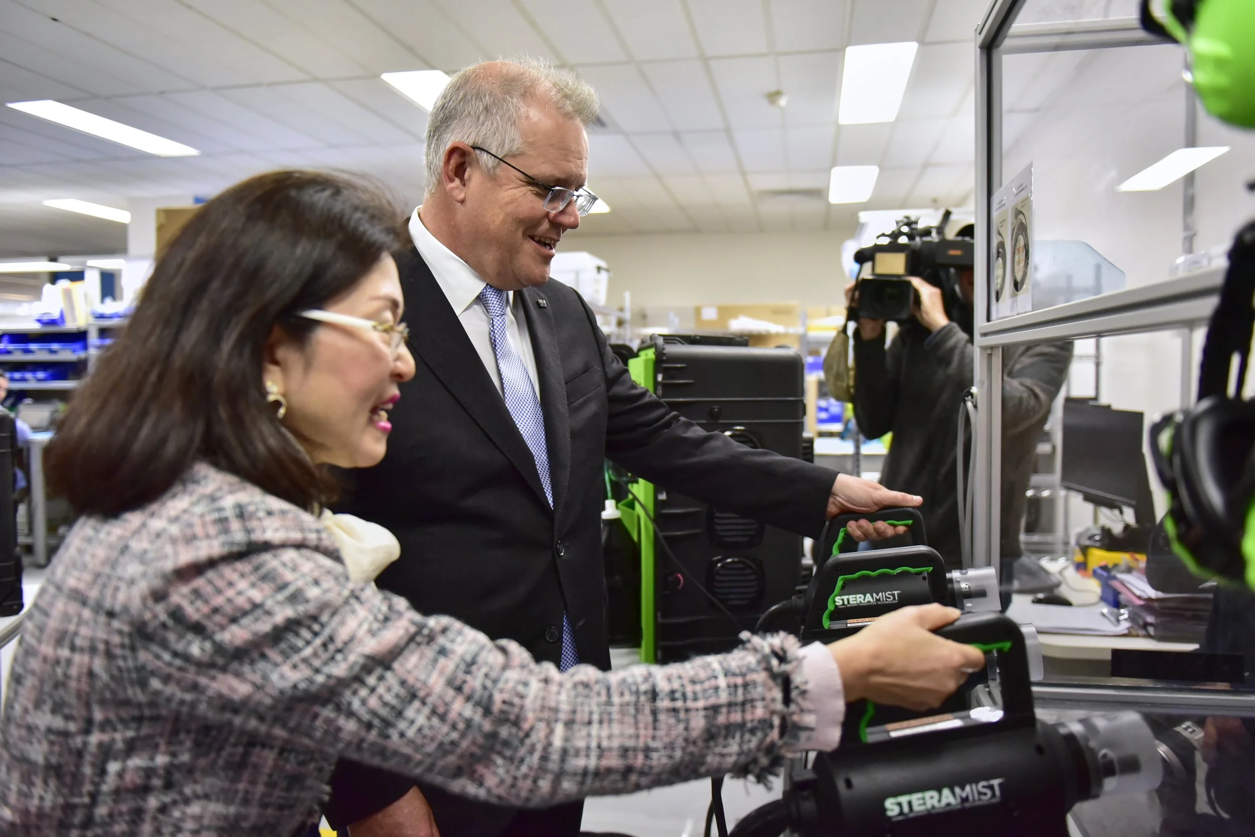 Two people, a woman and a man, working with sterilizer equipment in a lab or manufacturing environment, with a person operating a camera in the background.