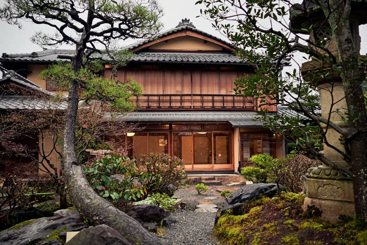 Traditional Japanese-style house with wooden exterior, surrounded by a Japanese garden with moss, rocks, and pruned trees.