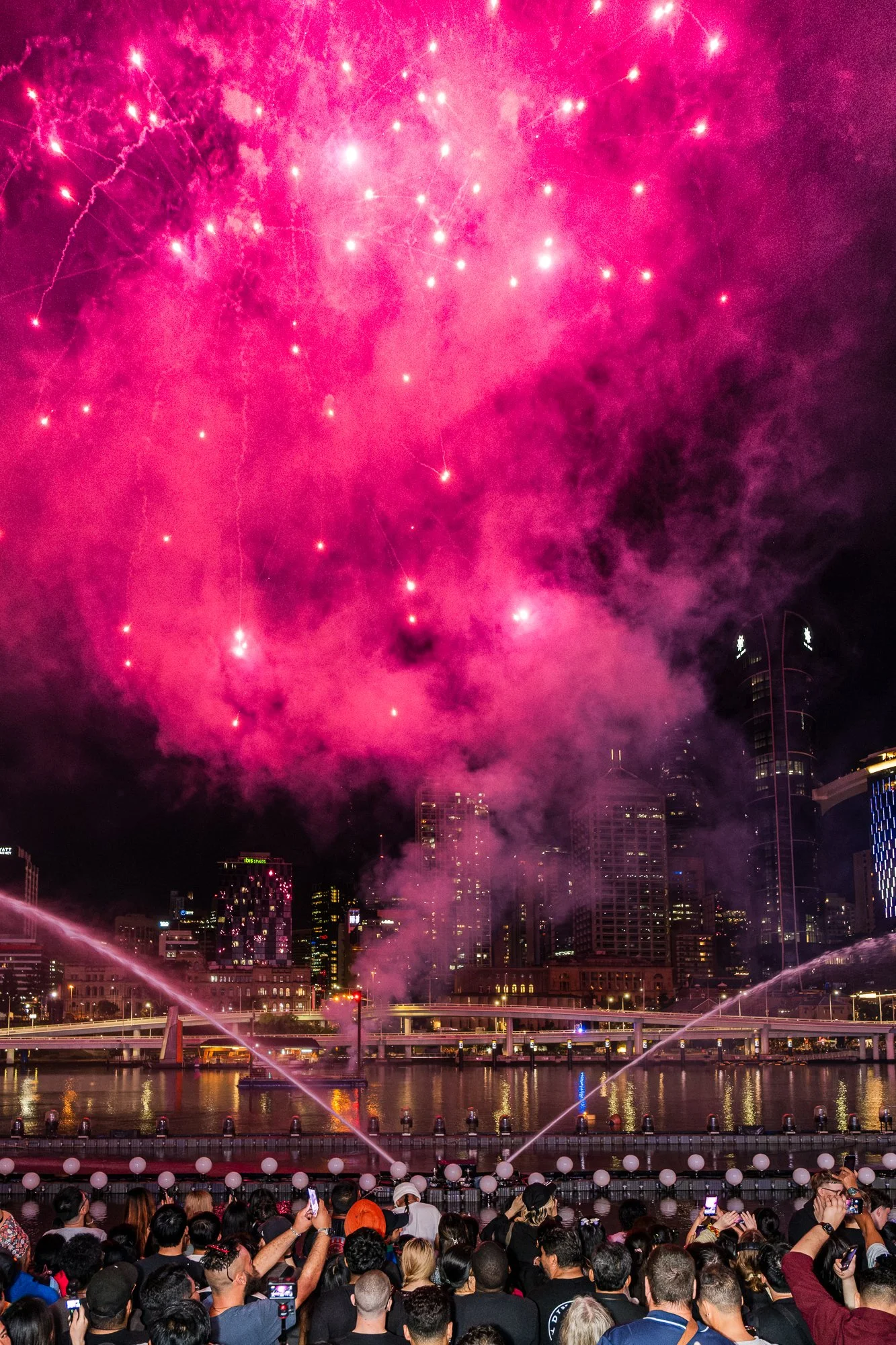 Crowd of people watching fireworks display over city skyline at night with pink and purple fireworks lighting up the sky.