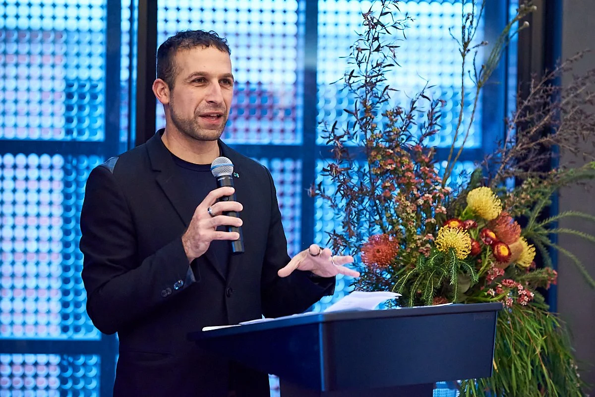 A man in a black blazer speaking into a microphone at a podium decorated with a large floral arrangement, with blue patterned paneling in the background.