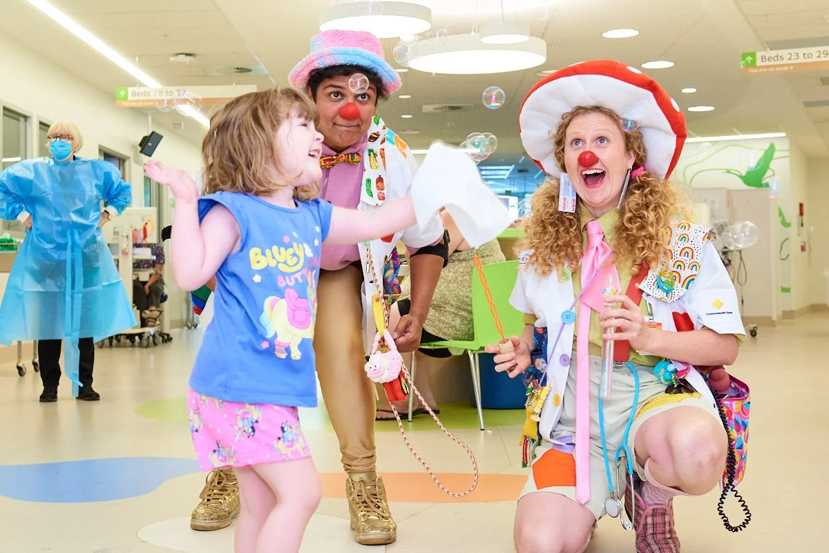 Children and clowns in a hospital, dressed as doctors and jesters, engaging in playful activities with colorful decorations and bubbles around.
