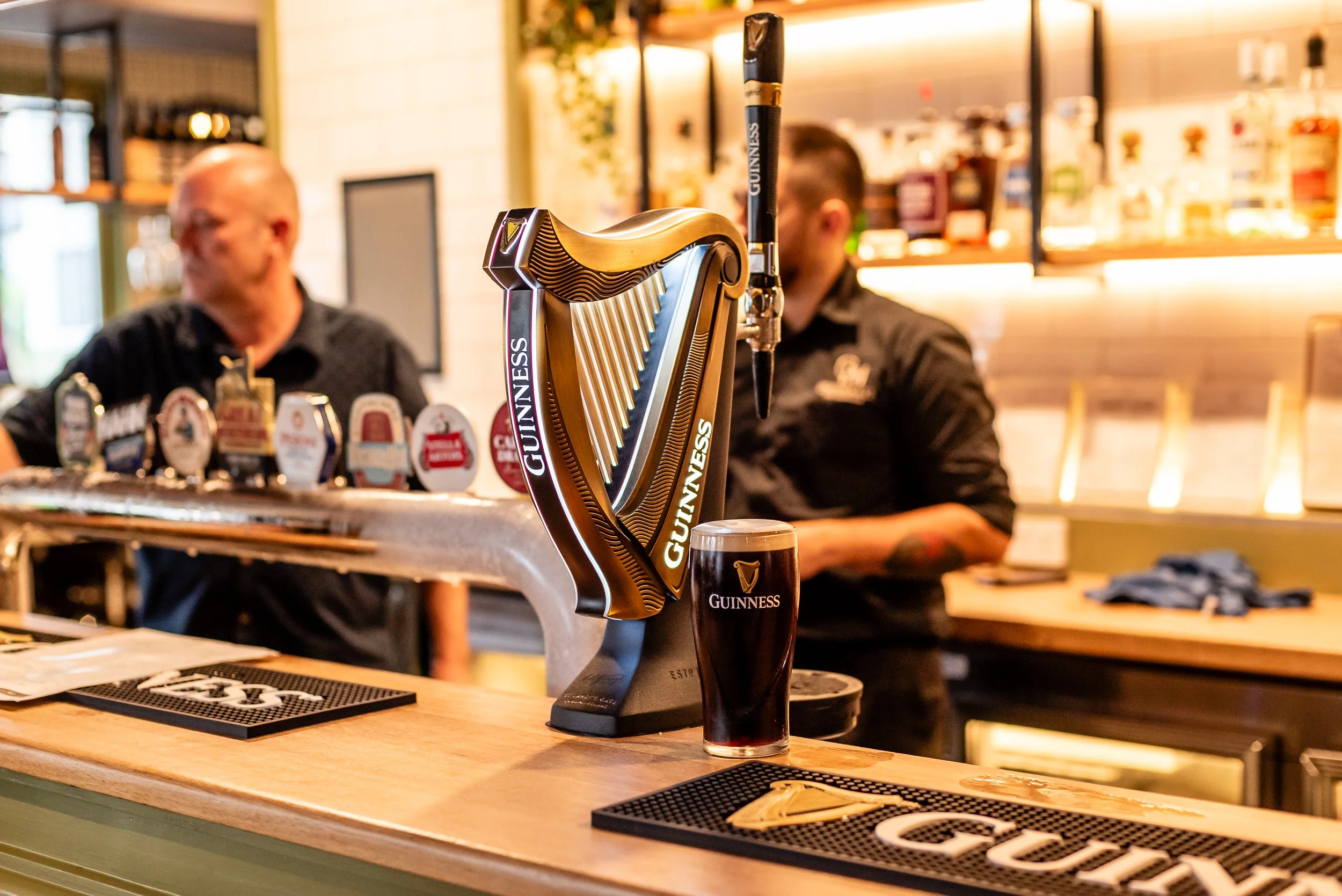 A Guinness harp beer tap in focus at a bar, with blurred patrons in the background, and a glass of Guinness pint on the bar counter.