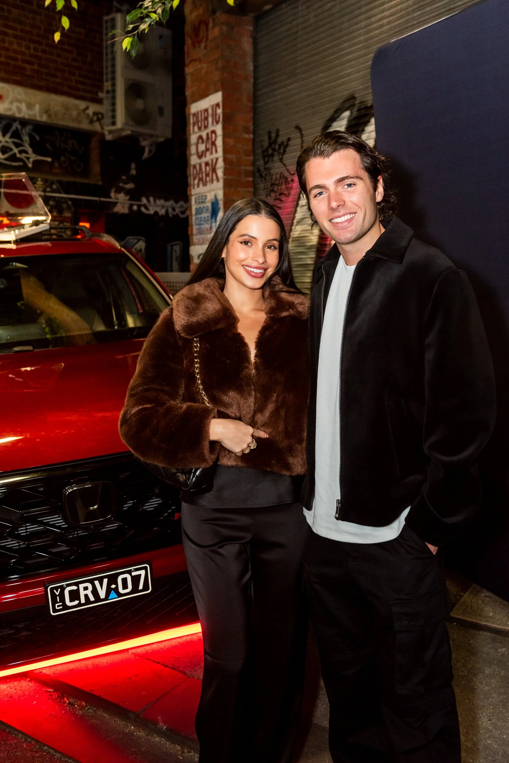A woman and a man are standing together outside at night in front of a red car with a license plate reading 'CRV-07'. The woman is wearing a brown faux fur coat, black pants, and has long dark hair, while the man is wearing a black jacket and black p
