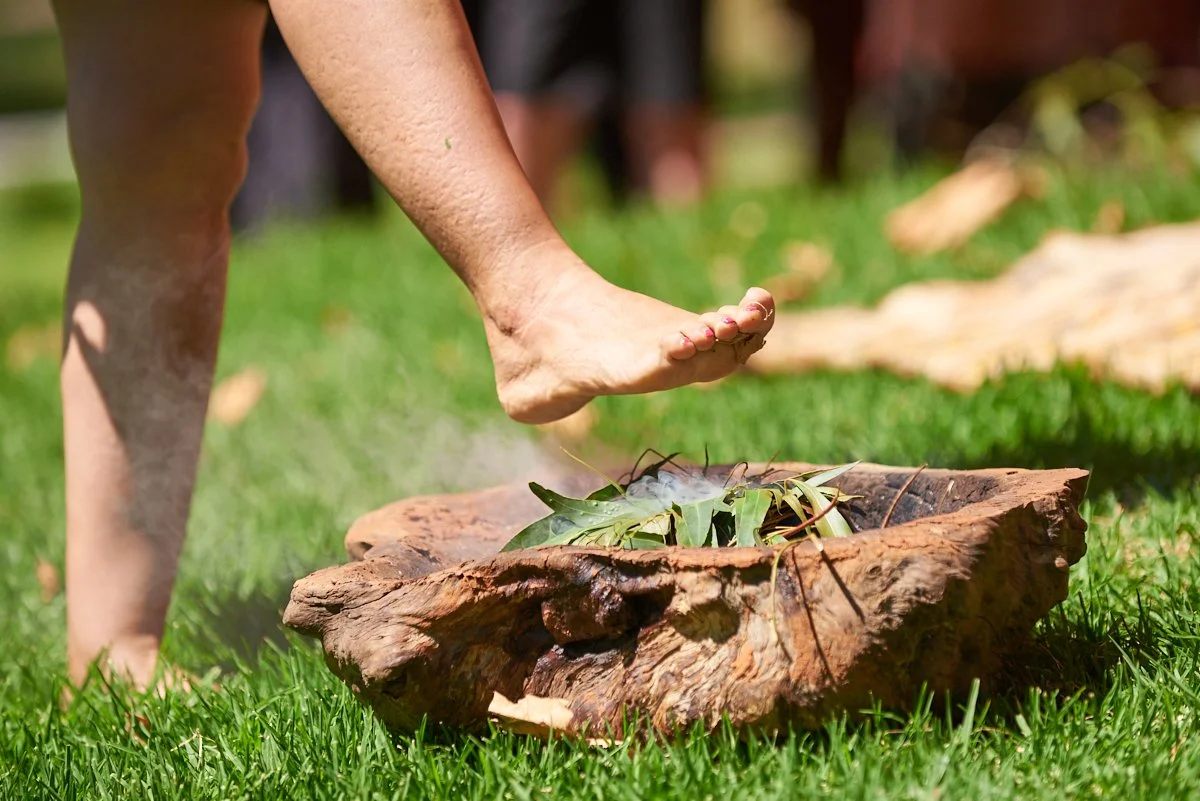 A person with dark skin is barefoot, balancing on one foot above a wooden platform with leaves and sticks, on a grassy field.