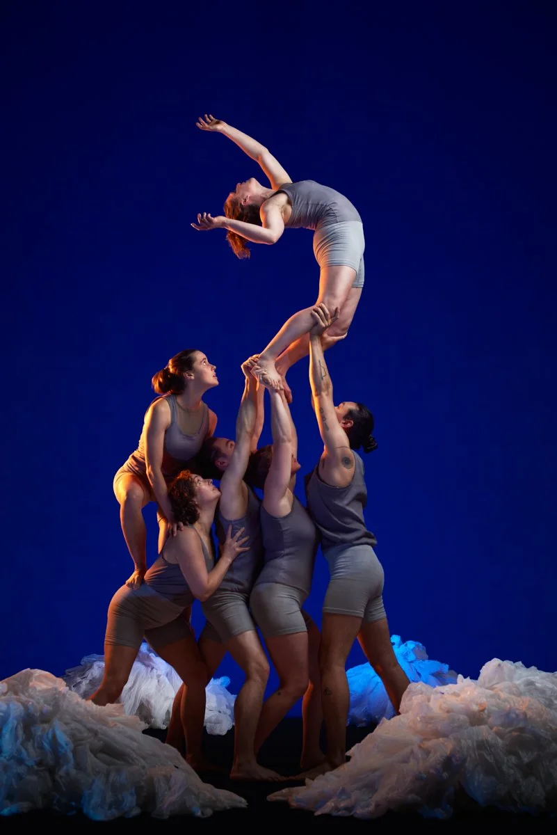 Five women performing an acrobatic pose on stage with cloud-like props, against a dark blue background.