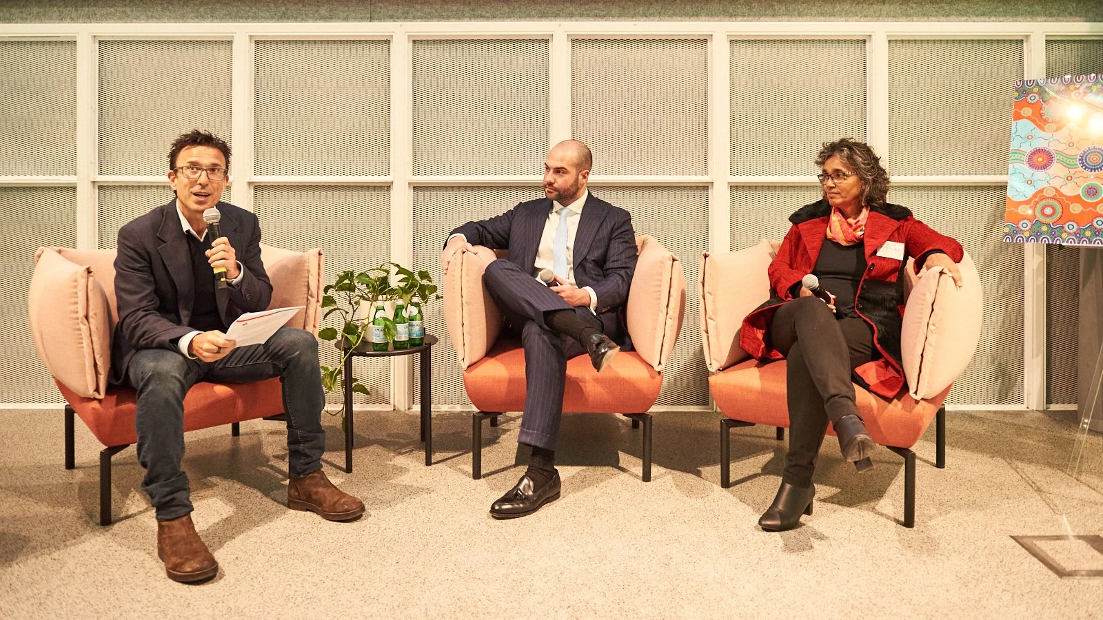 Three people sitting on pink armchairs in a panel discussion, with the man on the left holding a microphone and a paper, the man in the center with crossed legs holding a microphone, and the woman on the right holding a microphone and wearing a red c