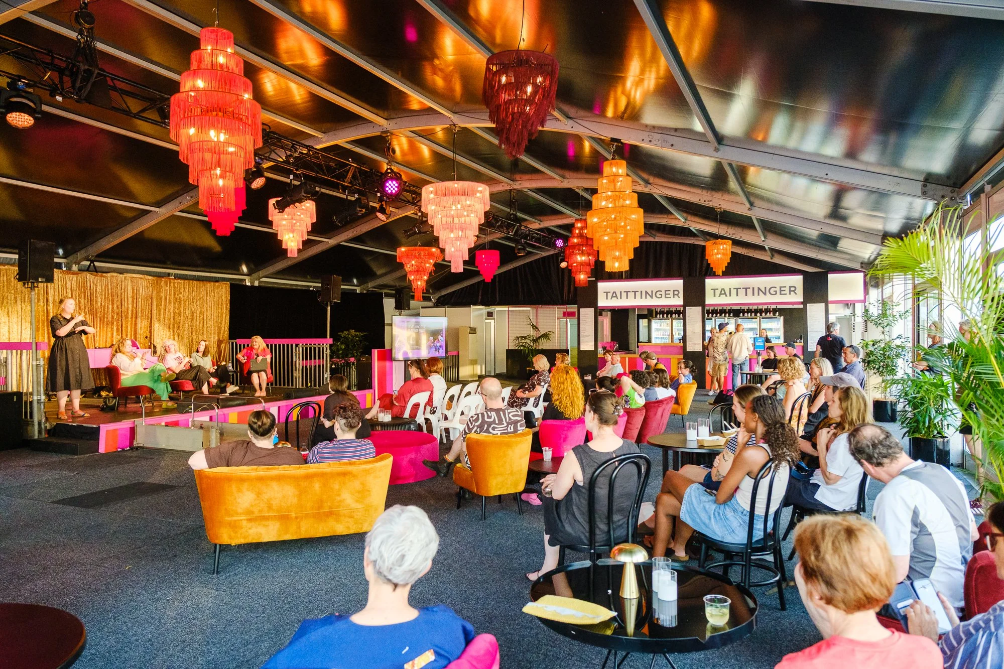 A lively indoor event with a stage where a woman is speaking to an audience seated in colorful chairs. The ceiling has large, vibrant red and orange chandeliers, and there are multiple screens and speakers behind the stage. The audience includes dive