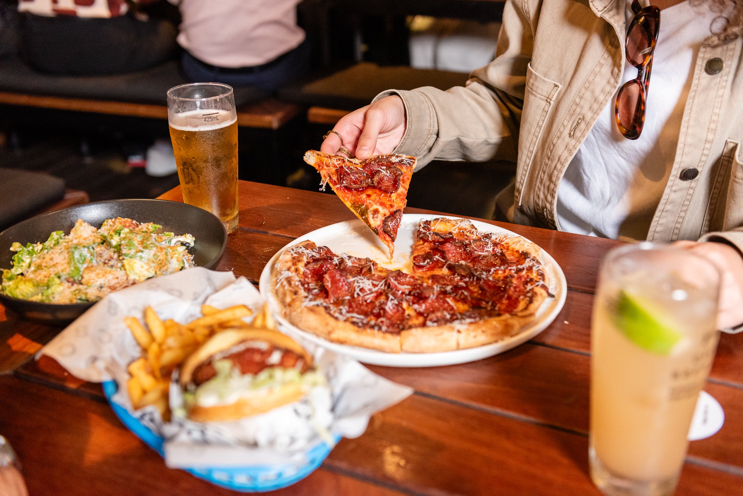 A person in a beige jacket with sunglasses hanging from the collar is holding a slice of pepperoni pizza over a white plate on a wooden table. There is a glass of beer, a salad, French fries, and other food items on the table.