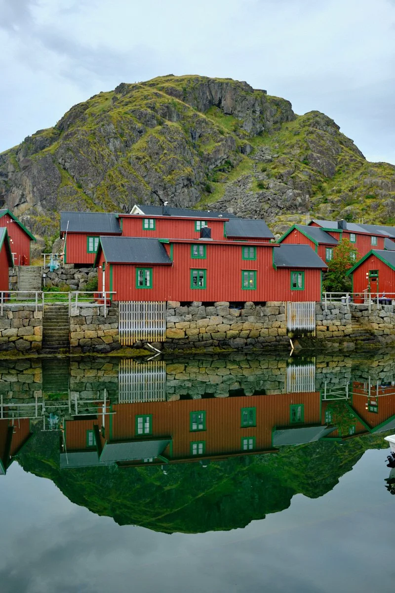 Red houses with green window frames along a waterfront, reflected in the calm water, with a large green mountain in the background under a cloudy sky.