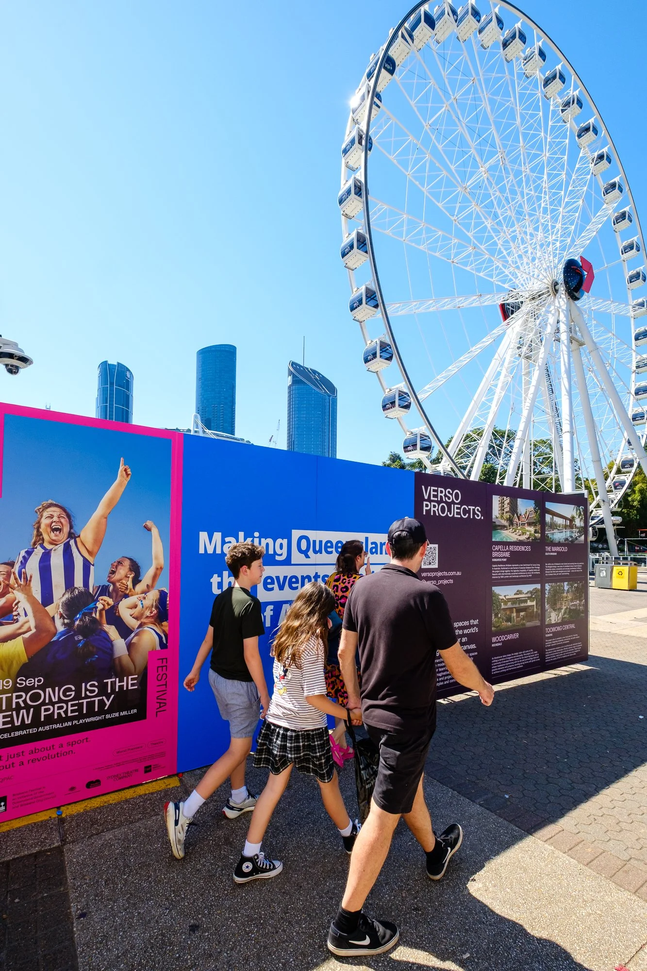 People walking past an outdoor advertisement display with aFerris wheel and modern skyscrapers in the background. The advertisements promote events and projects in Brisbane, Australia.