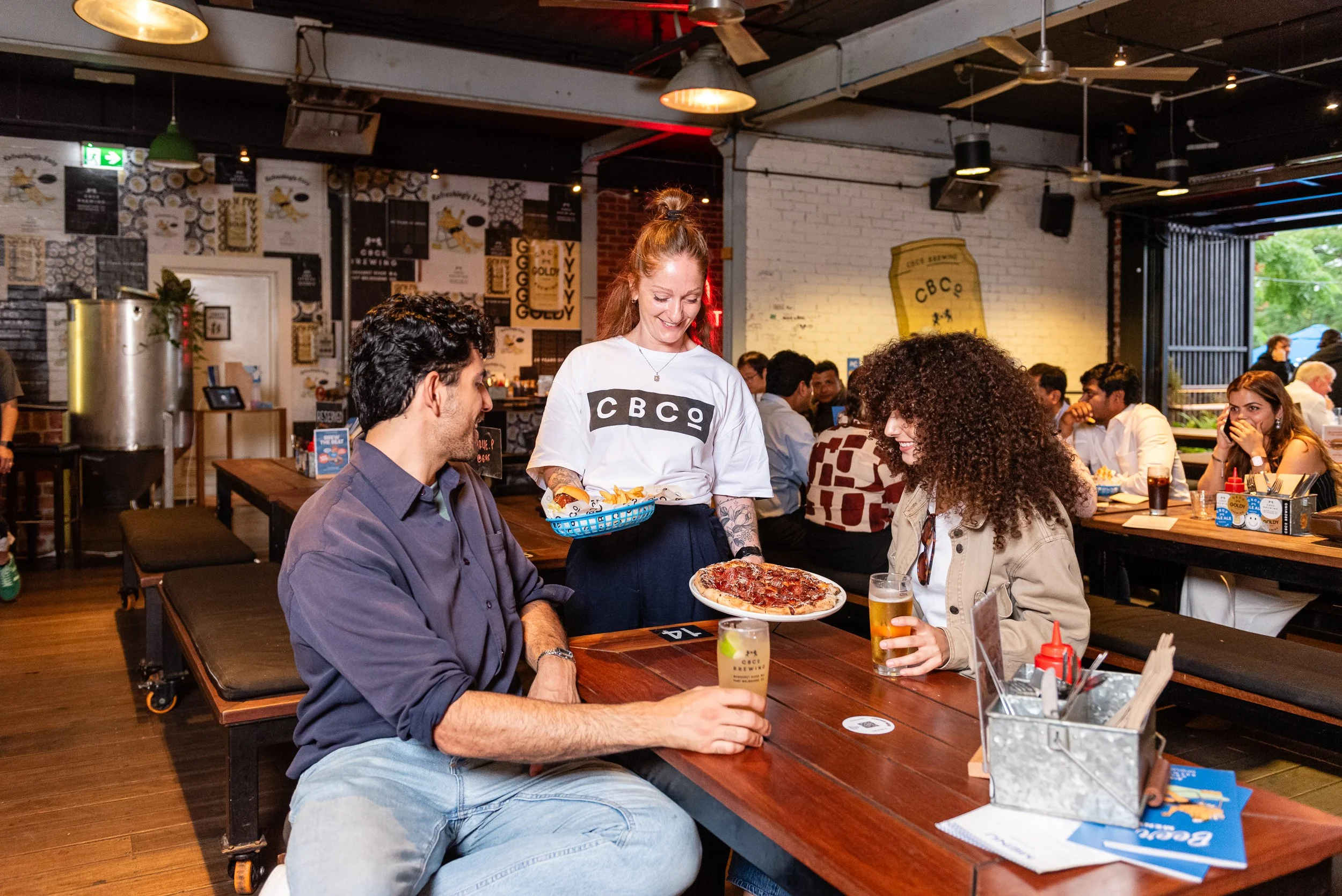 A group of people dining in a restaurant with a waitress serving pizza. The restaurant has wooden tables, chairs, and decorated walls with posters and art. The atmosphere is lively and casual.