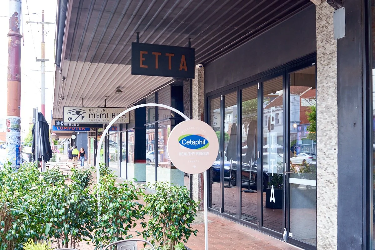 Outdoor shopping strip with storefronts, including Etta and Ancient Memories restaurant and cafe, and a Cetaphil sign in front, with plants and people walking along the sidewalk.