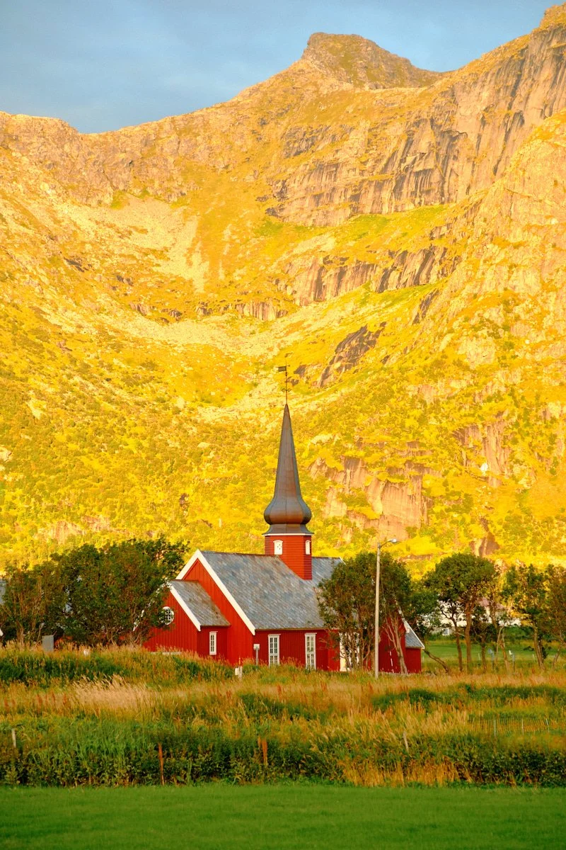A red church with a tall black steeple is situated in a green field, with mountains covered in yellow and green foliage in the background under a clear blue sky.