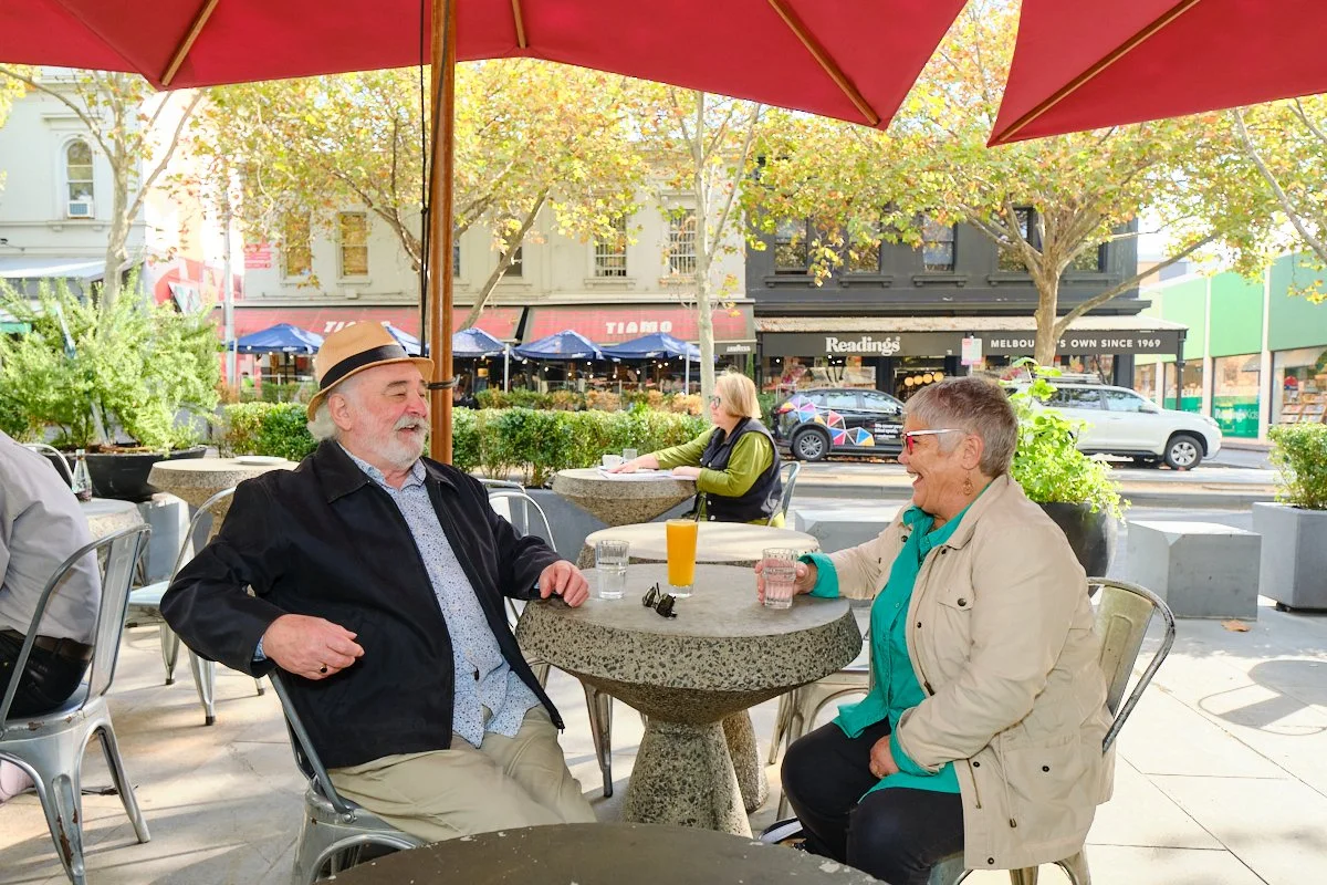 Two elderly people, a man with a beige hat and a woman with short gray hair, sitting at an outdoor cafe table under a large red umbrella, smiling and talking to each other, with drinks on the table. Trees and storefronts are visible in the background
