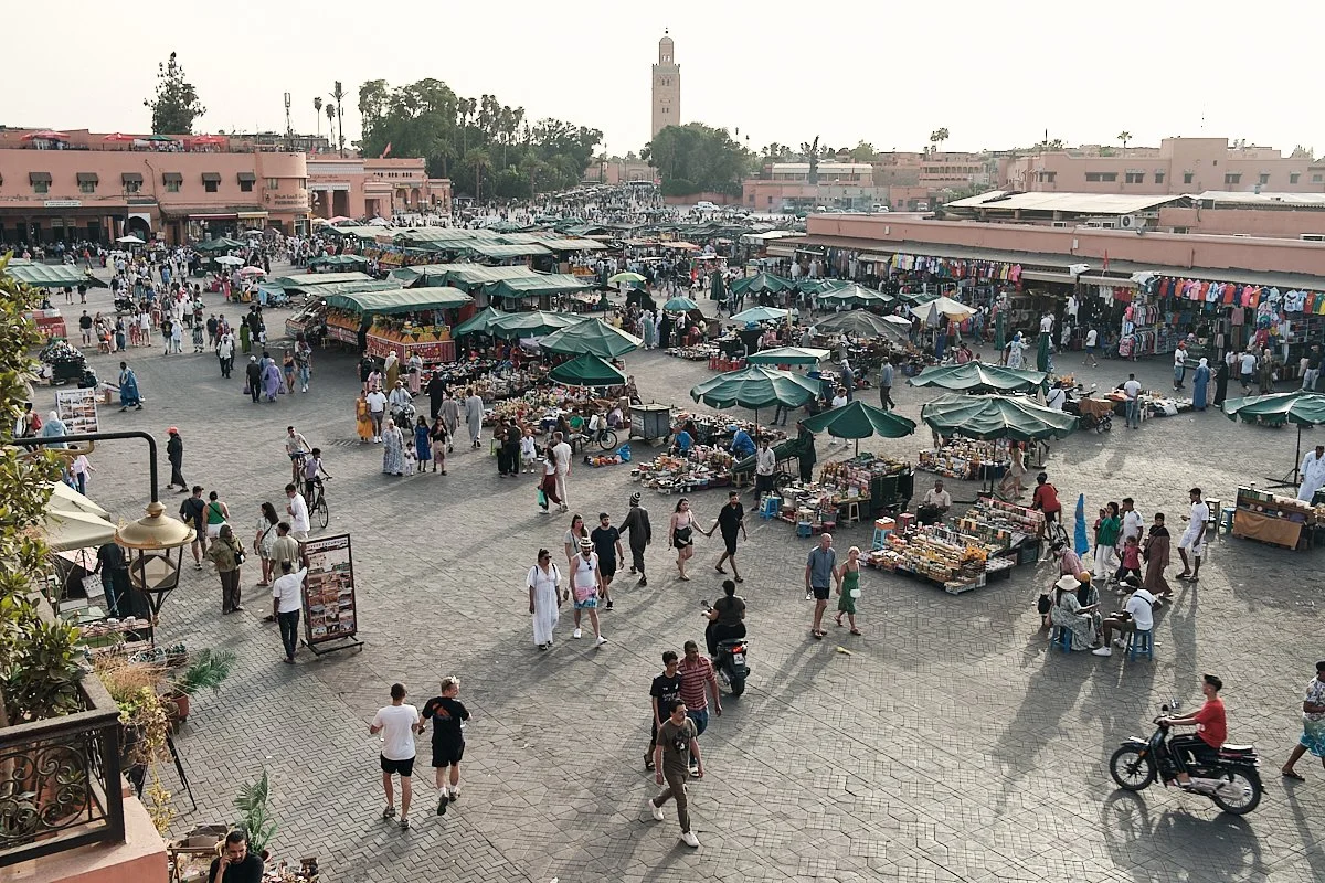 Open-air market with numerous vendors selling goods under green umbrellas, people walking and shopping in a large paved square with pink buildings and a tall clock tower in the background.