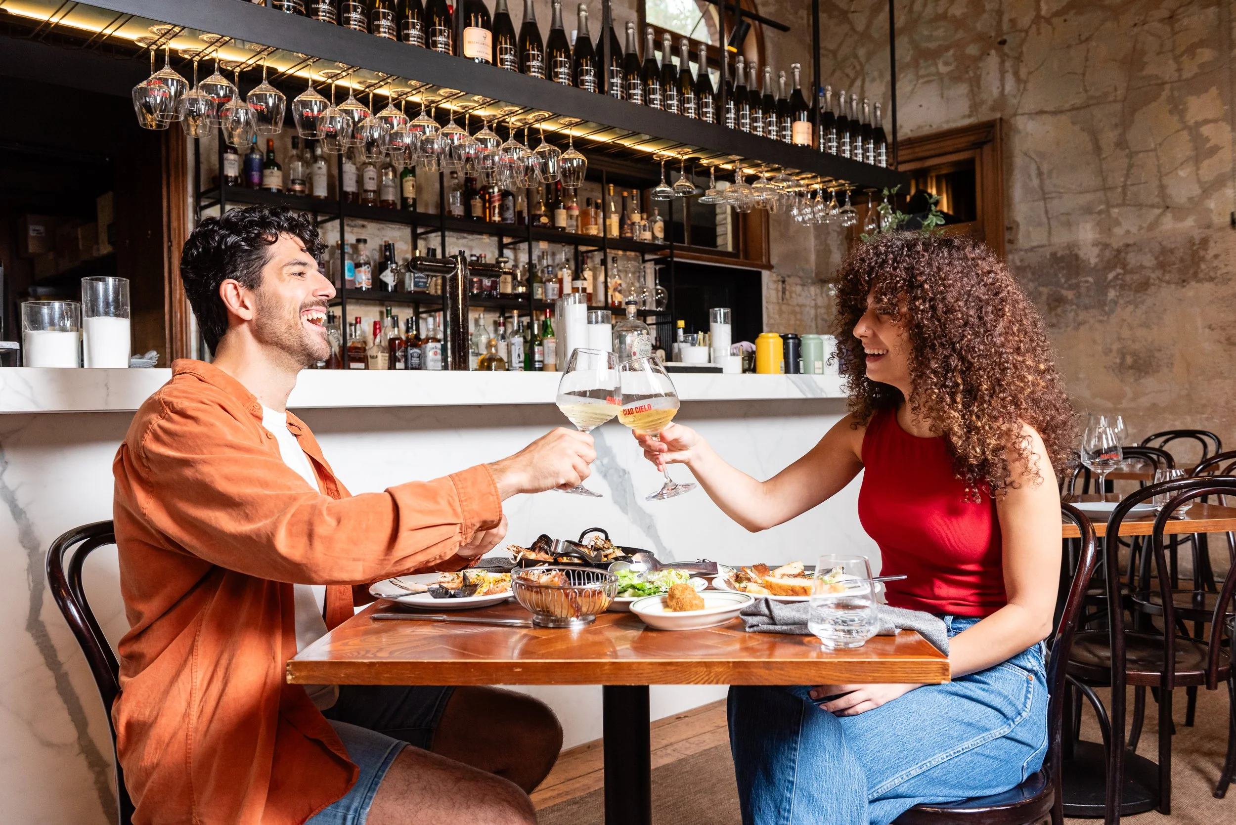 A man and woman sitting at a wooden table in a restaurant, clinking wine glasses and smiling. The table has plates of food, and the background features a bar with bottles and wine glasses hanging upside down.