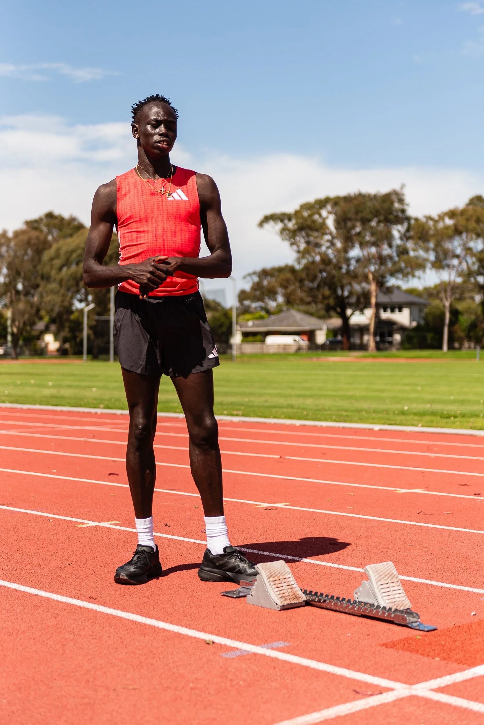 A male athlete standing on a running track, preparing to race, wearing a red sleeveless shirt, black shorts, and black running shoes, with a starting block in front of him under a bright sky with trees and buildings in the background.