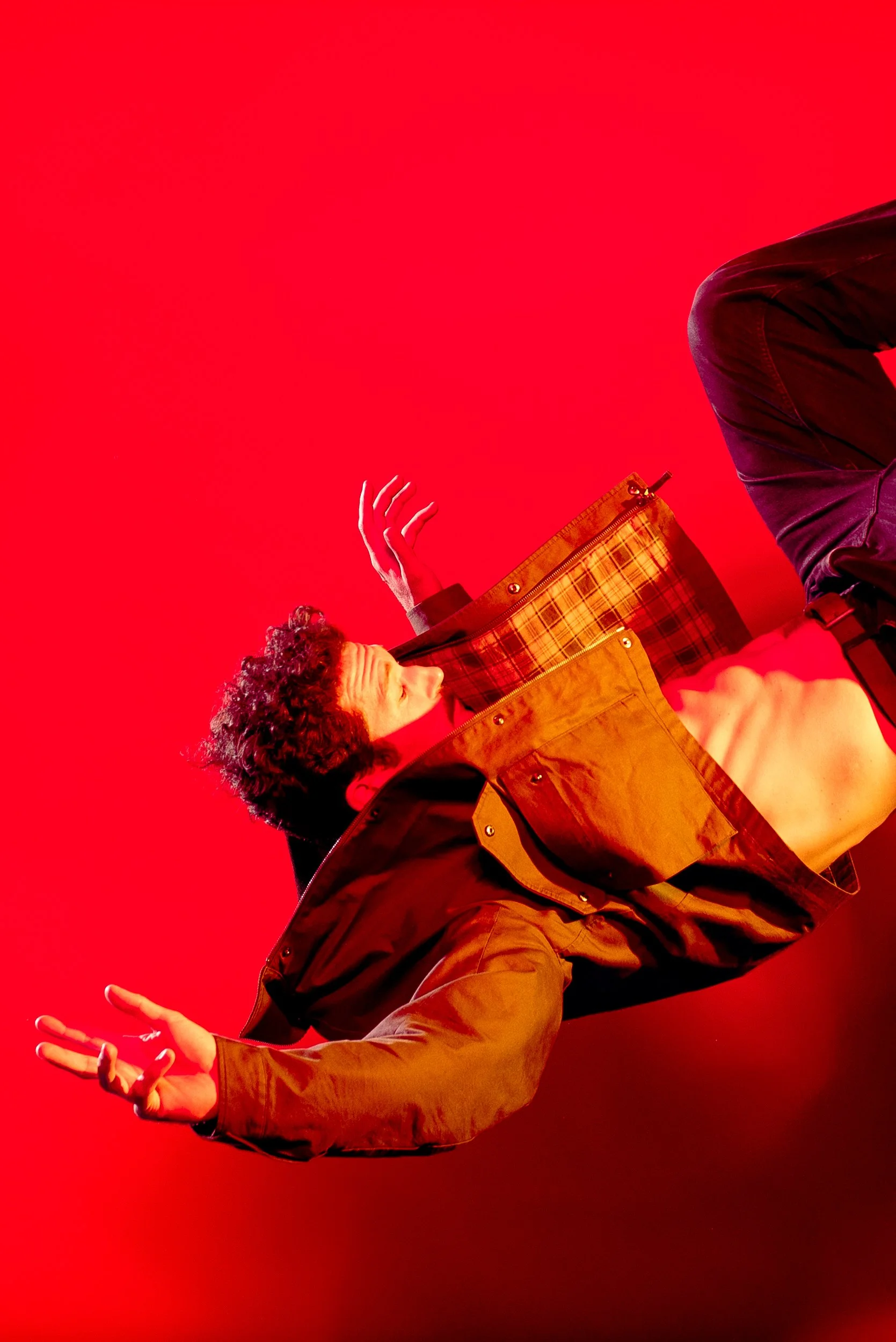 A person with curly hair wearing an open brown jacket, exposing their bare chest, poses against a bright red background. They have one hand raised and the other extended outward.
