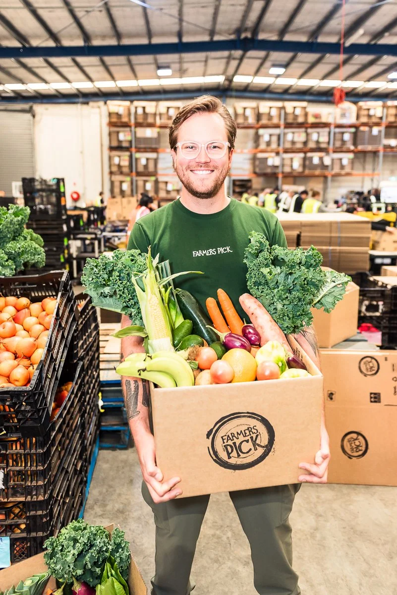 A smiling man wearing glasses and a green shirt that says 'Farmers Pick' is holding a large box of fresh vegetables at a warehouse or grocery facility. The box contains kale, zucchini, carrots, and various other vegetables. There are more produce crates around him and warehouse shelves in the background.