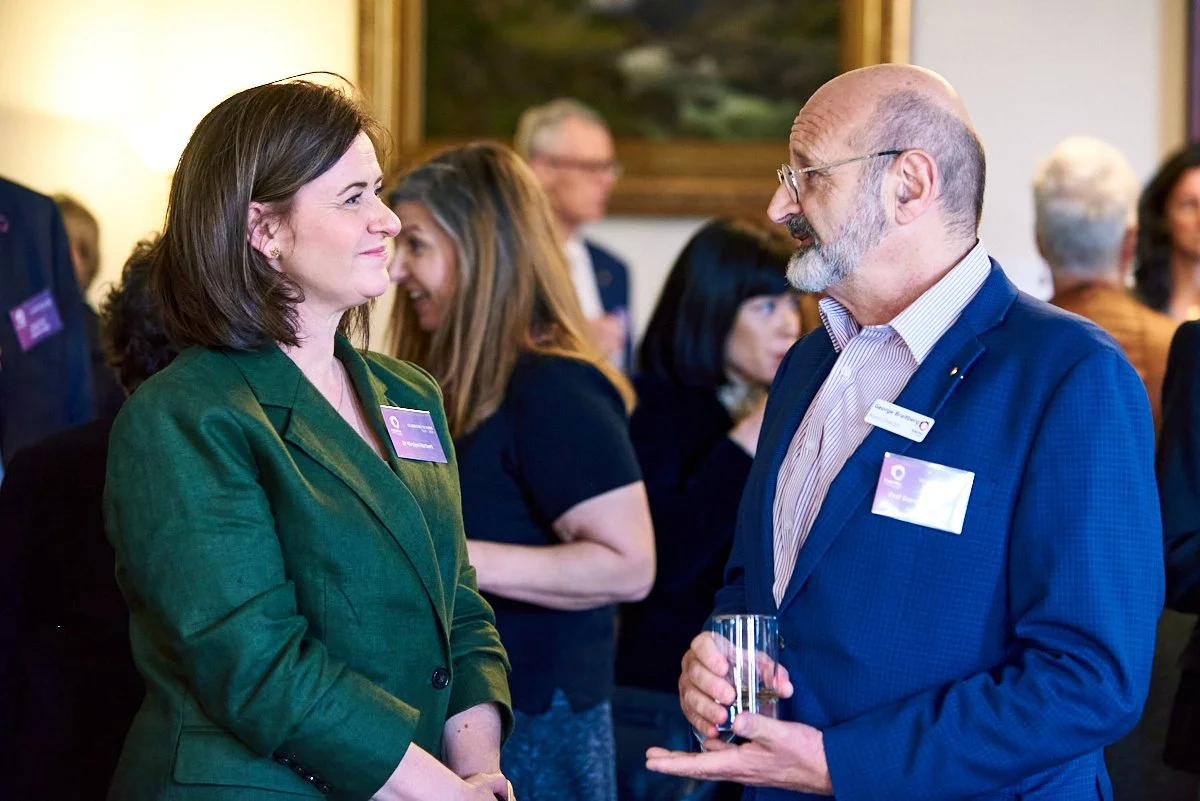 Two people, a woman in a green blazer and a man in a blue suit, are talking at a professional event. They are smiling and holding drinks. Other people are visible in the background.