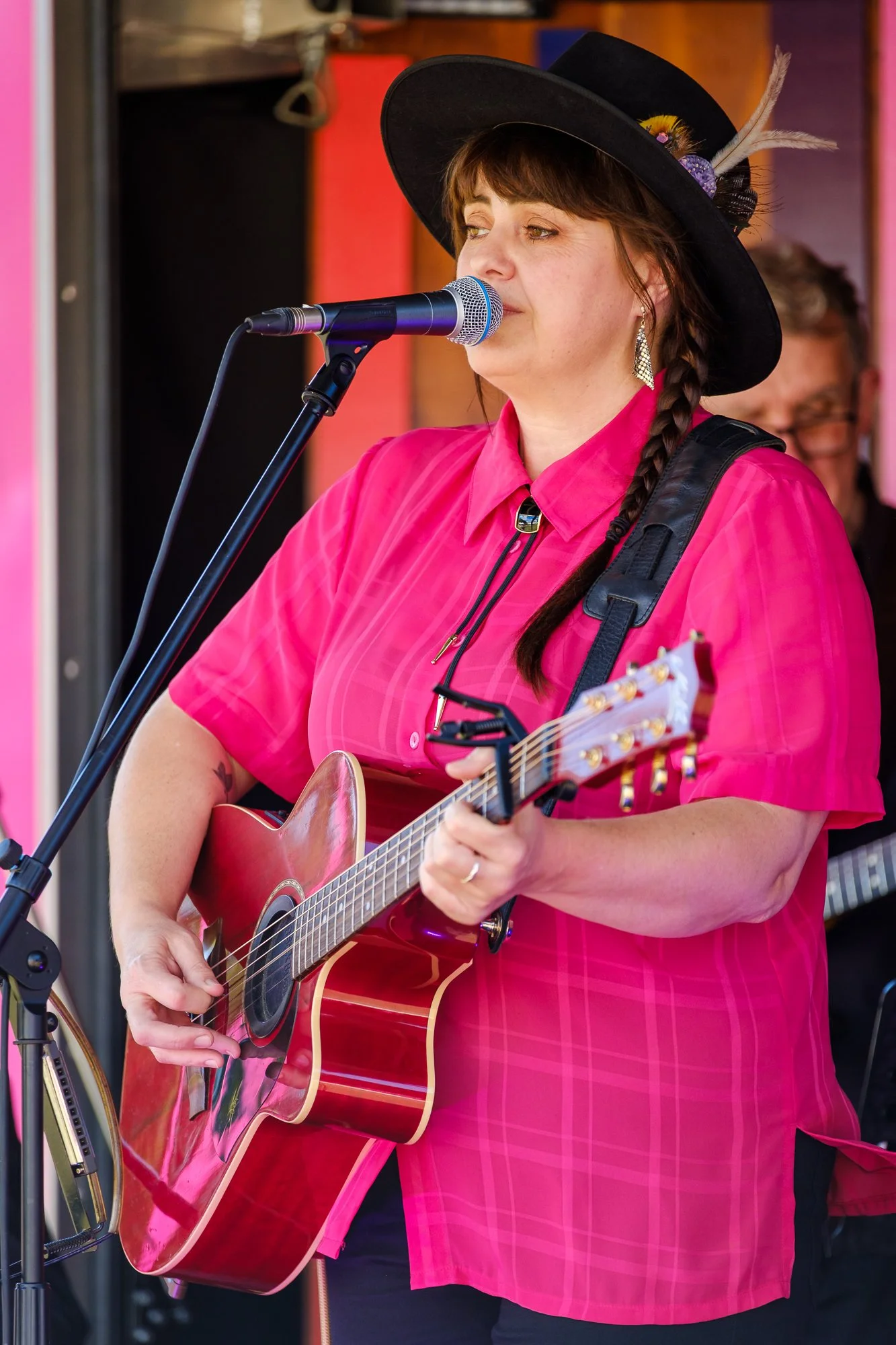 A woman wearing a pink shirt and a black wide-brimmed hat decorated with feathers, singing into a microphone while playing an acoustic guitar during a street performance.
