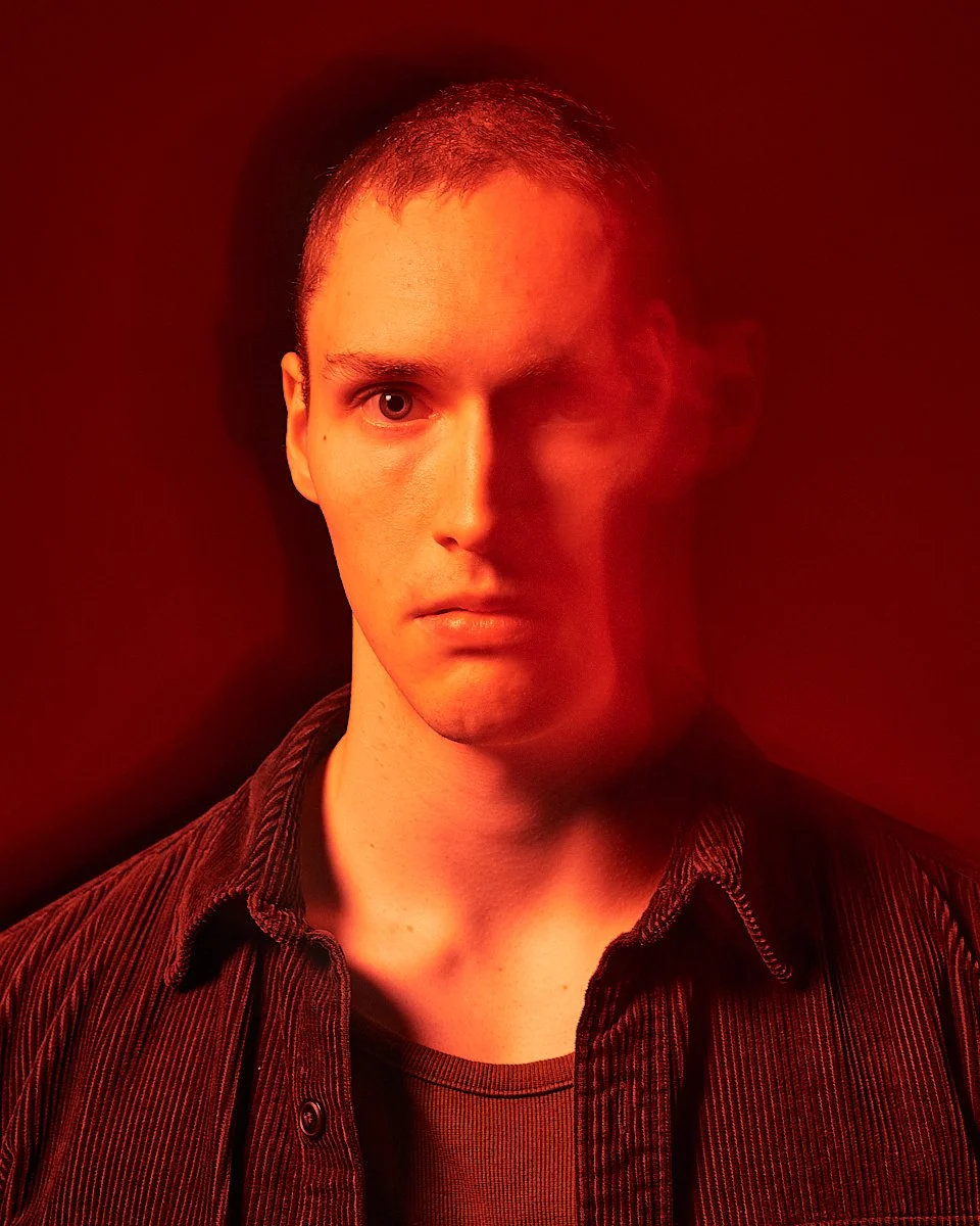 A young man with short hair and a serious expression, lit with red and orange lighting, wearing a dark shirt and jacket, against a dark background with a motion blur effect.