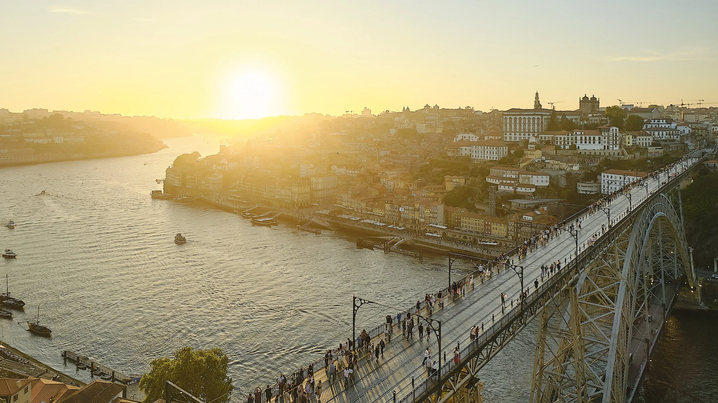 Dusk over a river with boats, a city with colorful buildings on a hillside, and a bridge filled with pedestrians.