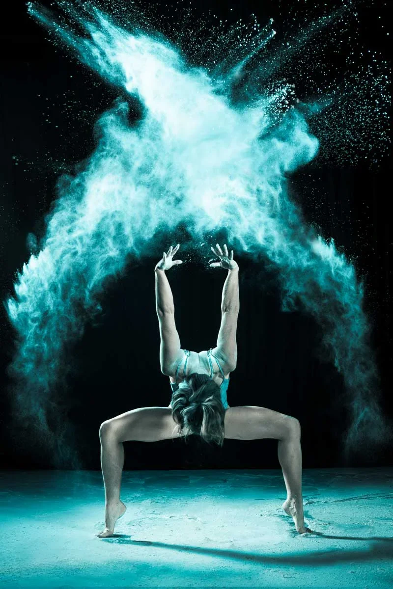 A woman performs a contorted yoga pose with water or powder exploding behind her, creating a dynamic display of motion and energy.