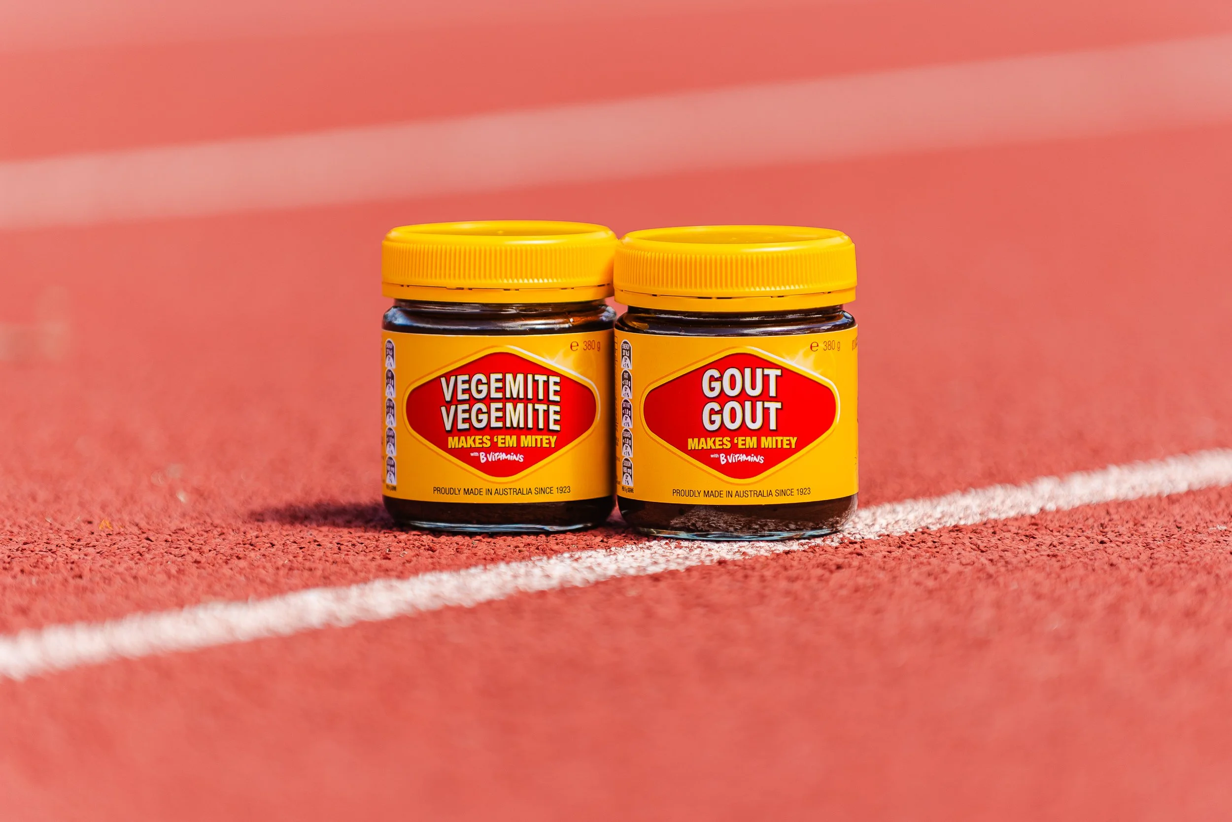Two jars of supplements on a red running track, labeled 'Vegemite Vegemite' and 'Gout Gout' with bright yellow lids and labels.