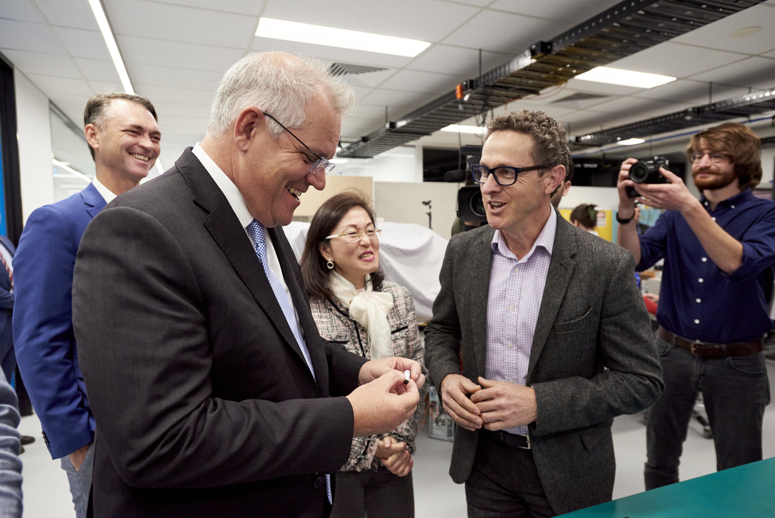 A group of five people engaged in conversation in an office or lab setting, with one person taking photos.