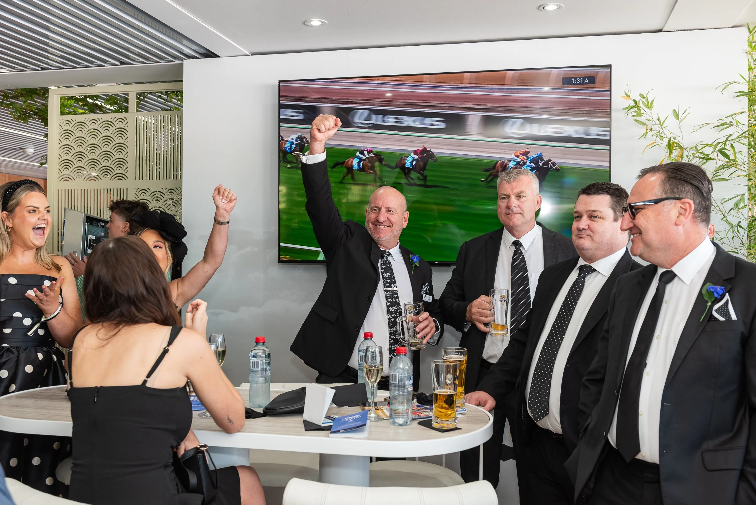 A group of people celebrating a horse race victory at a social gathering, with some raising their glasses and others smiling, in front of a large television screen showing a horse race.